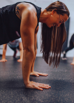 Woman in a black tank top doing a push-up on a gym floor, with others exercising in the background of a fitness class.